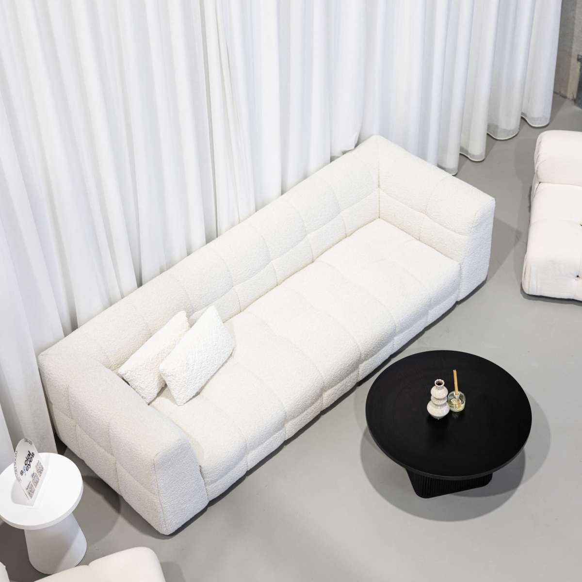 A white, tufted sofa is positioned against a backdrop of white curtains, accompanied by a circular black coffee table and a small white side table in a minimalist setting.