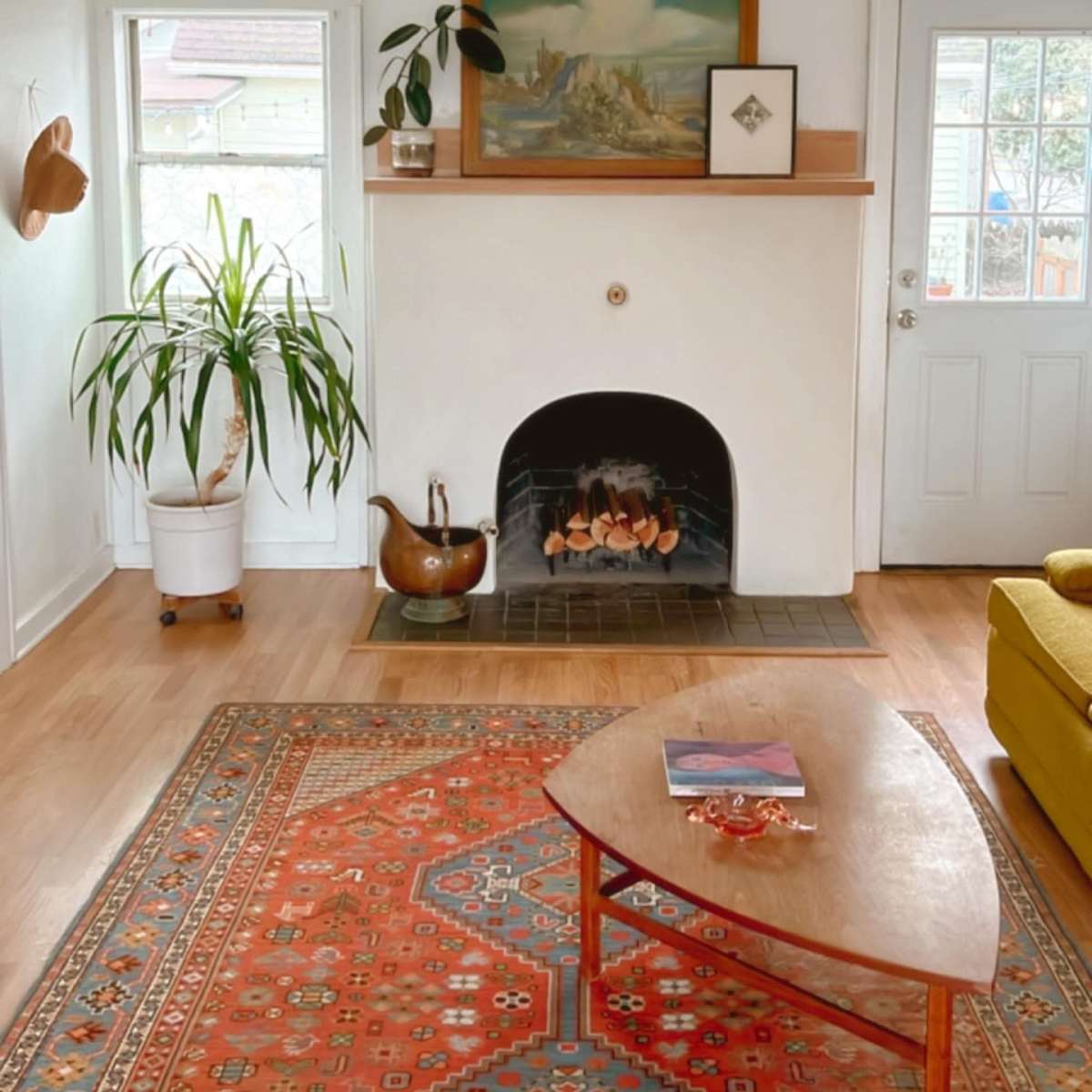 The scene features a cozy living room with a fireplace, a potted plant, a wooden coffee table, and a yellow sofa, accented by an area rug and artwork on the walls.