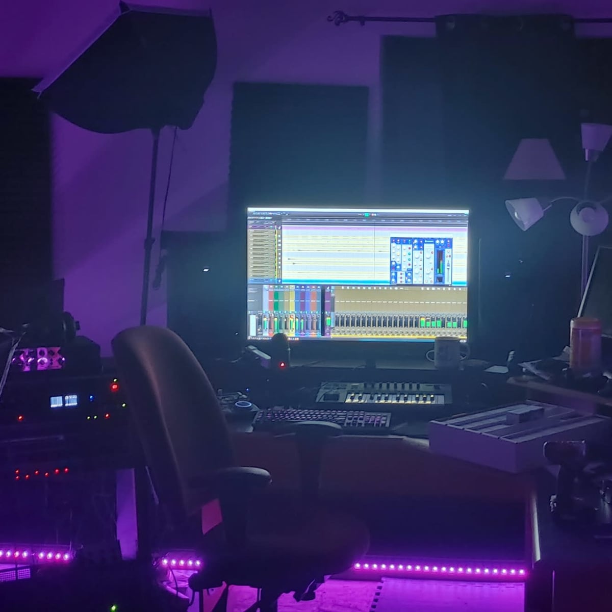 A well-equipped audio production studio features a large computer screen displaying mixing software, surrounded by various musical instruments and equipment, with ambient purple lighting.