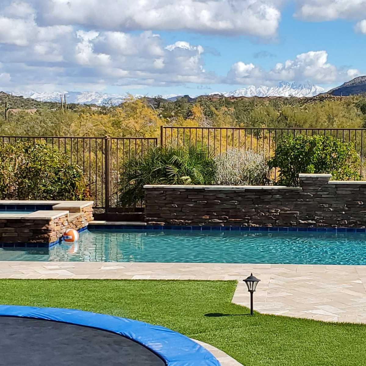 A dog rests on a grassy area next to a swimming pool, with mountains and blue skies in the background.