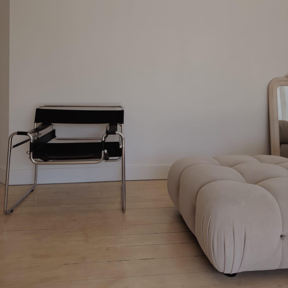 A black and chrome chair is positioned beside a beige tufted ottoman in a room with light wooden flooring and a plain wall.
