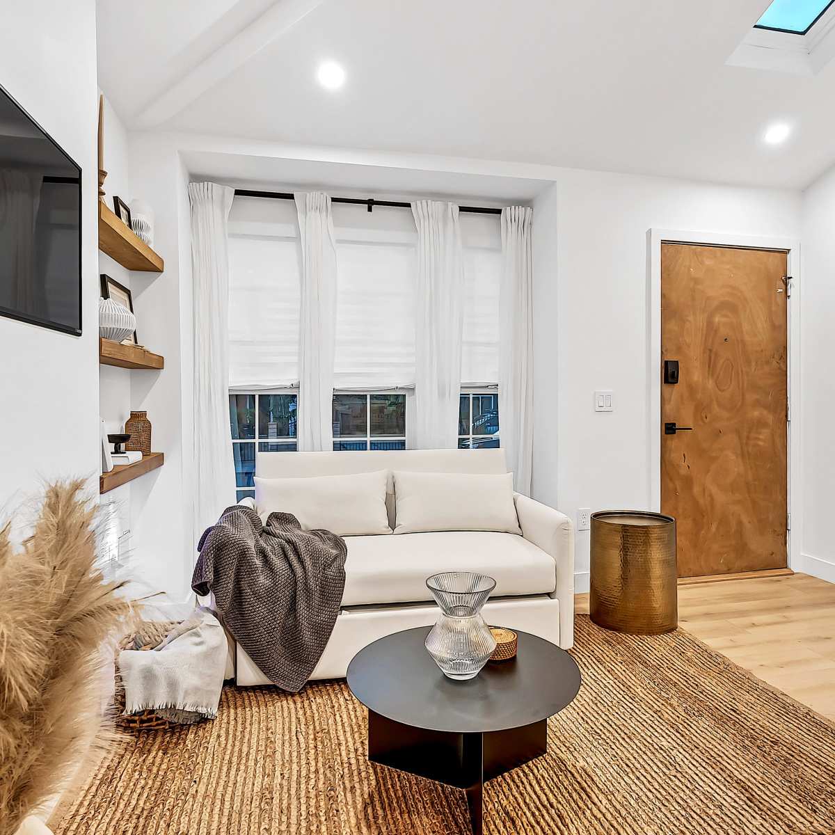 A modern living room featuring a white sofa, a round black coffee table, a textured area rug, and a wooden door with a glass panel.