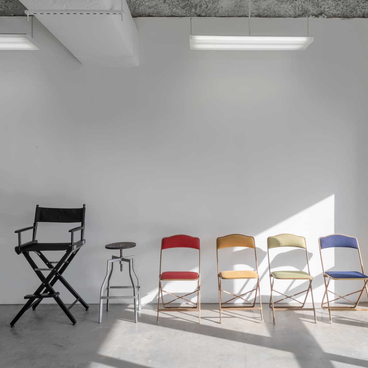 A black director's chair is positioned next to a metal stool and five colorful folding chairs against a white wall in a well-lit room.