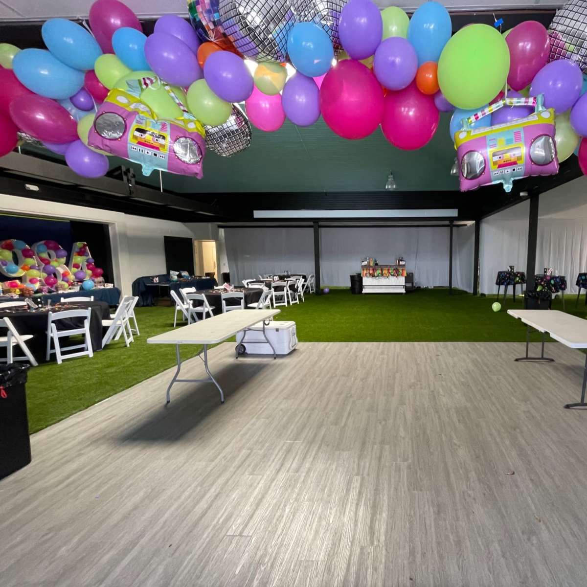 The venue features a decorated space with colorful balloons overhead, tables arranged for an event, and a grassy floor area.