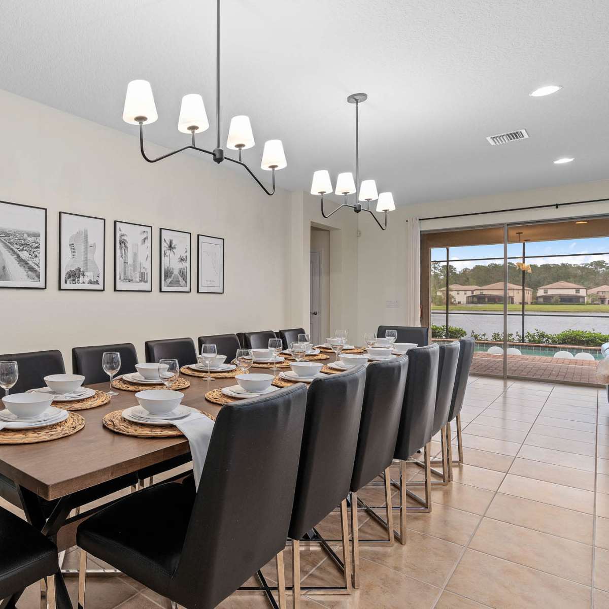 A long dining table set for a meal is displayed in a bright dining area with framed black-and-white photos on the wall and sliding glass doors leading to an outdoor space.