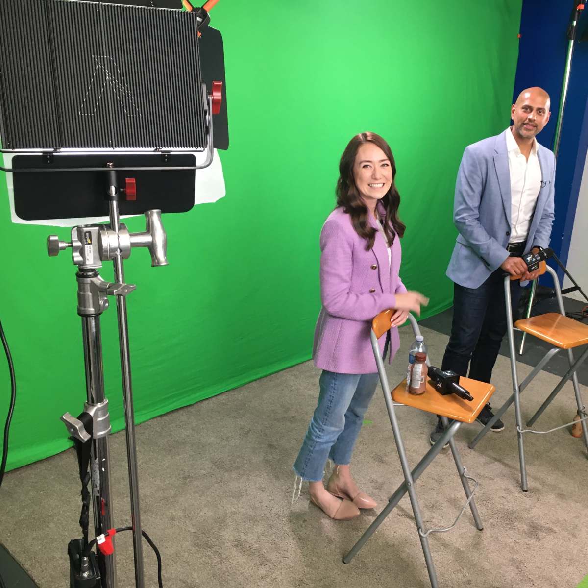 Two individuals stand on a green screen set, one in a purple blazer and the other in a light blue jacket, with chairs and equipment visible around them.