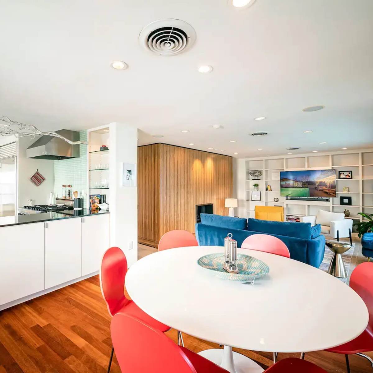 A modern kitchen and living area feature an open layout with a round dining table and red chairs adjacent to a blue sofa and a wooden accent wall.