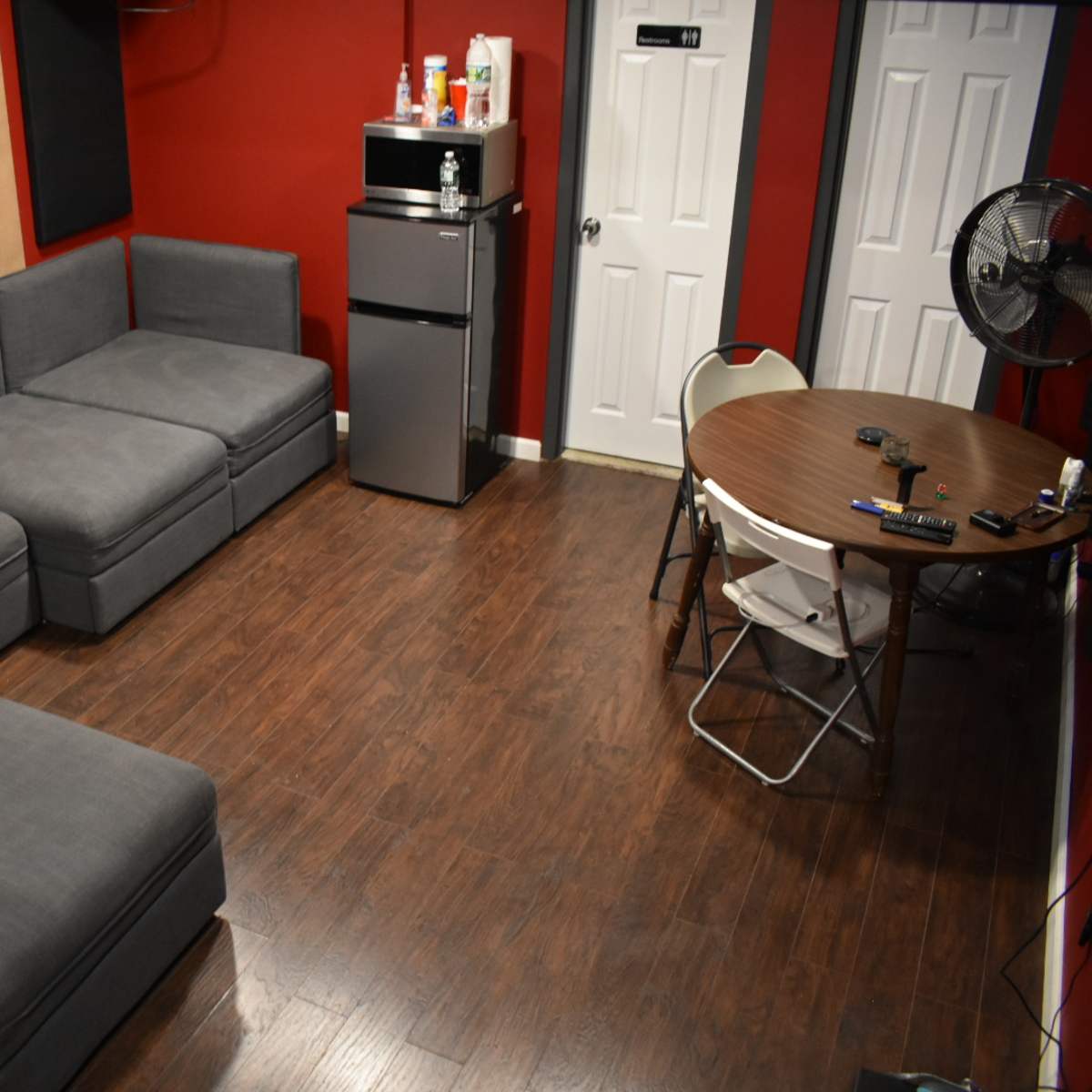 A small room with red walls, featuring a gray sectional sofa, a round table with chairs, and a mini refrigerator in one corner.