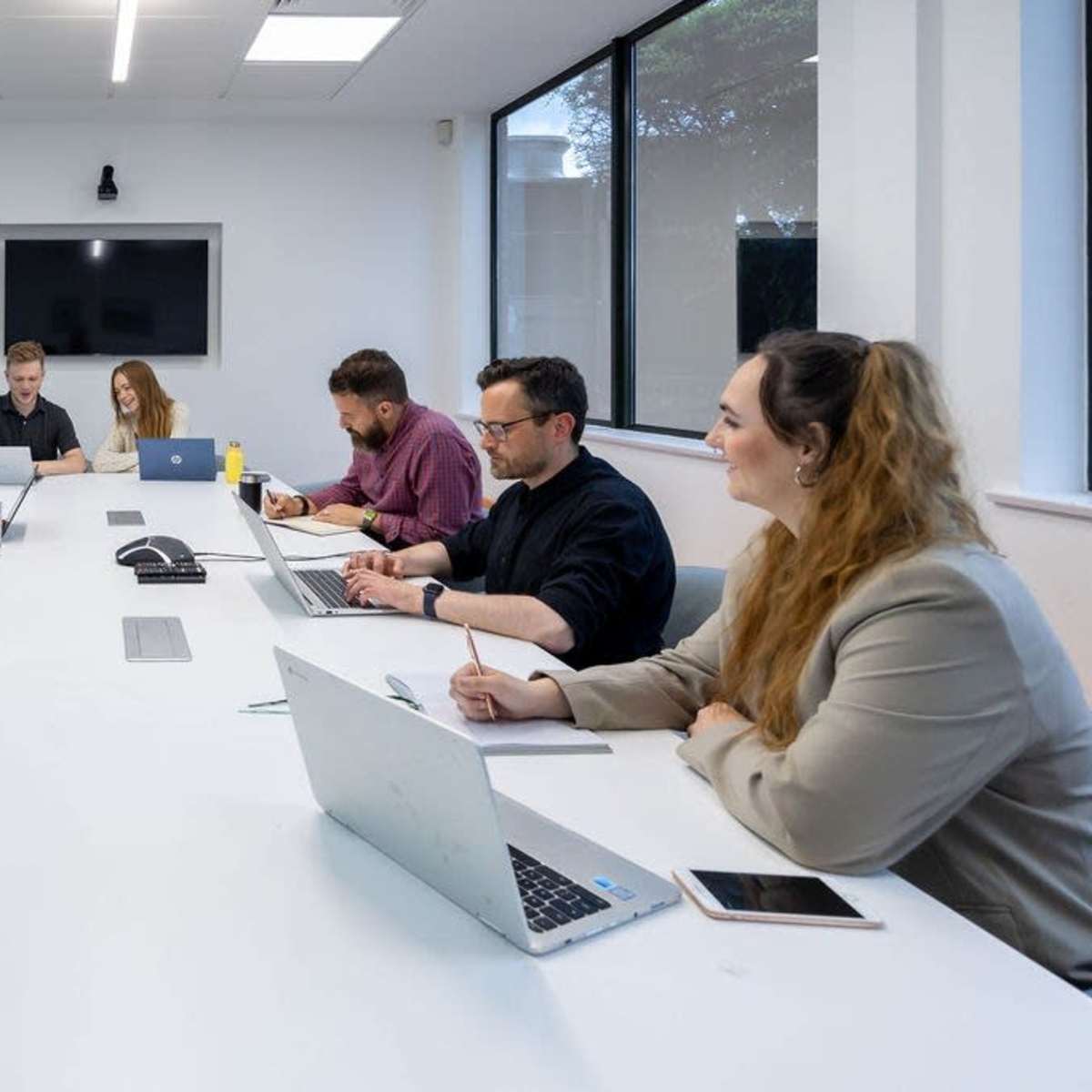 A group of six people is seated around a long white table in a conference room, engaged in discussions and working on laptops.