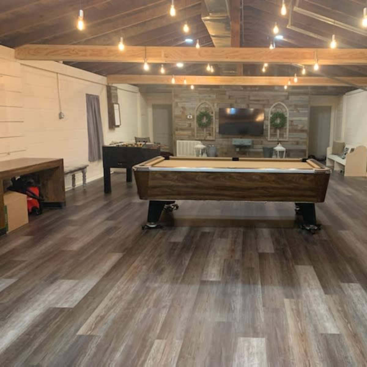 A spacious indoor room with a pool table in the center, wooden beams on the ceiling, and a television mounted on the wall.