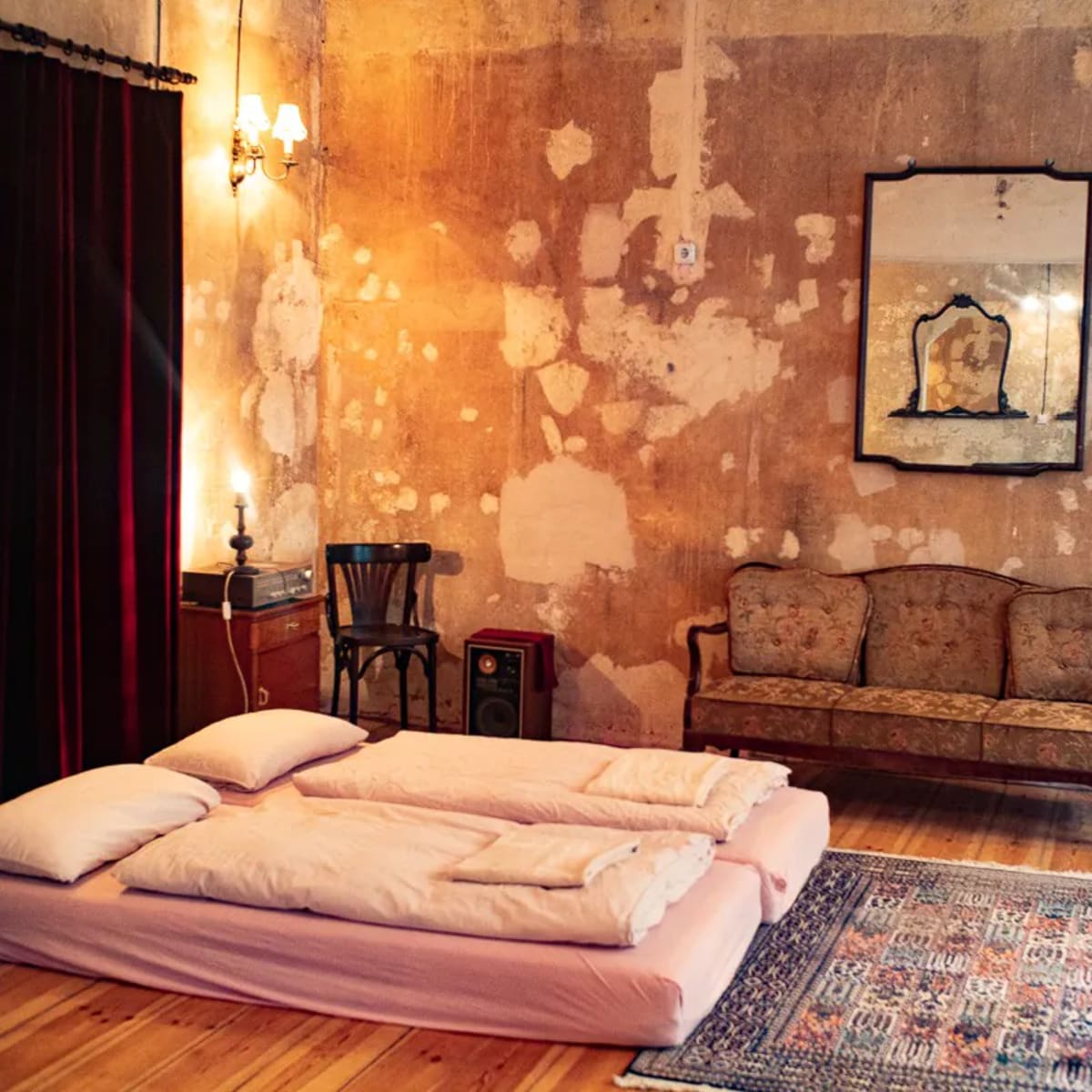 A sparsely furnished room with a simple bedding arrangement on the floor, a vintage sofa, a side table, and a patterned rug on a wooden floor, all set against a textured, peeling wall.