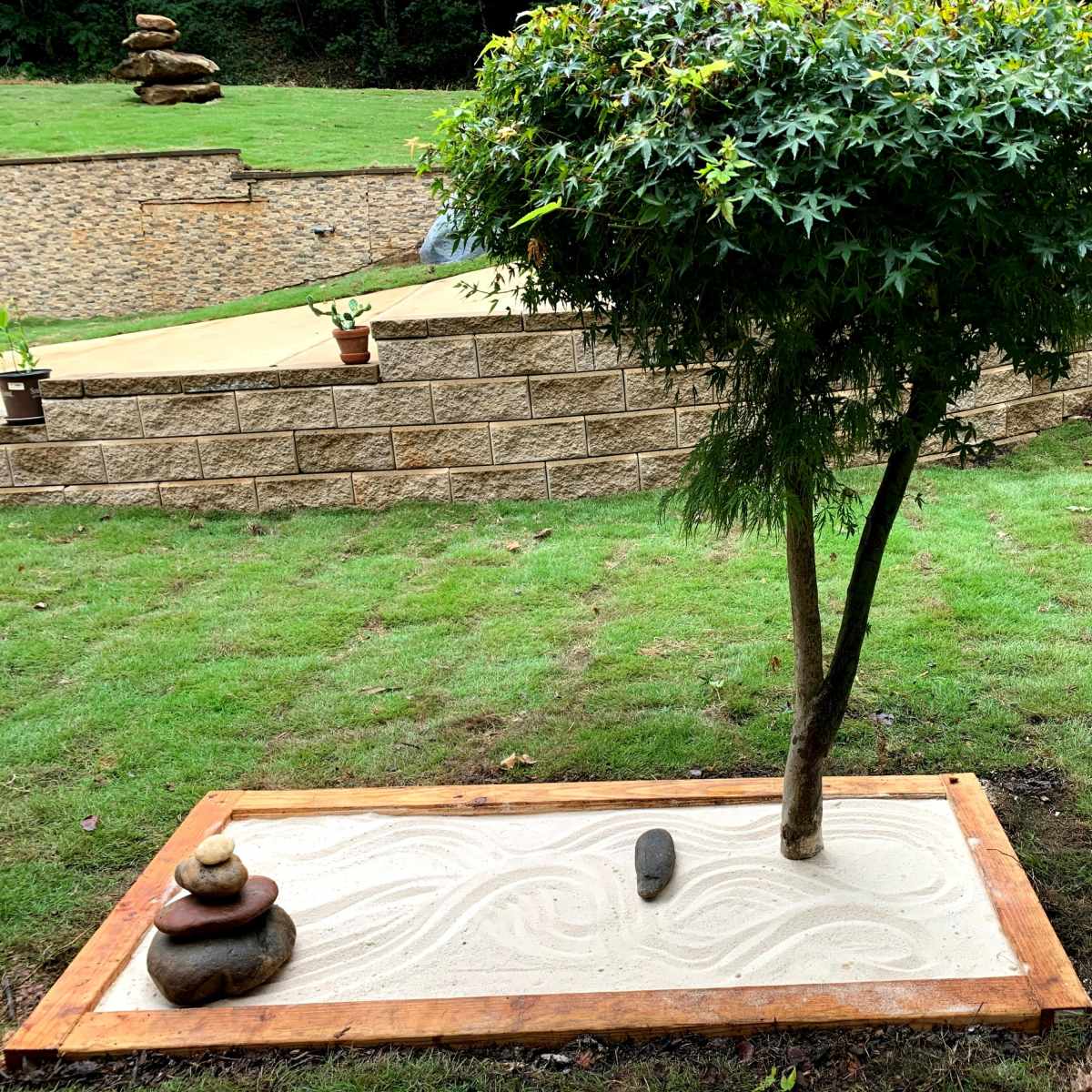 A small tree stands next to a sand garden framed by wooden borders, with stacked stones arranged on one side.