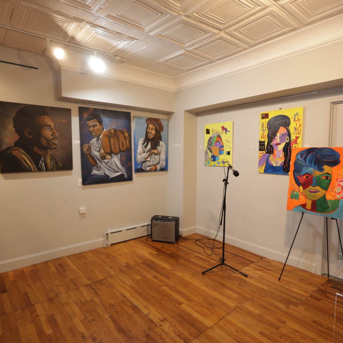 An art gallery featuring several paintings on the walls, including portraits and a colorful artwork on an easel, with a microphone positioned in front of a blank wall.