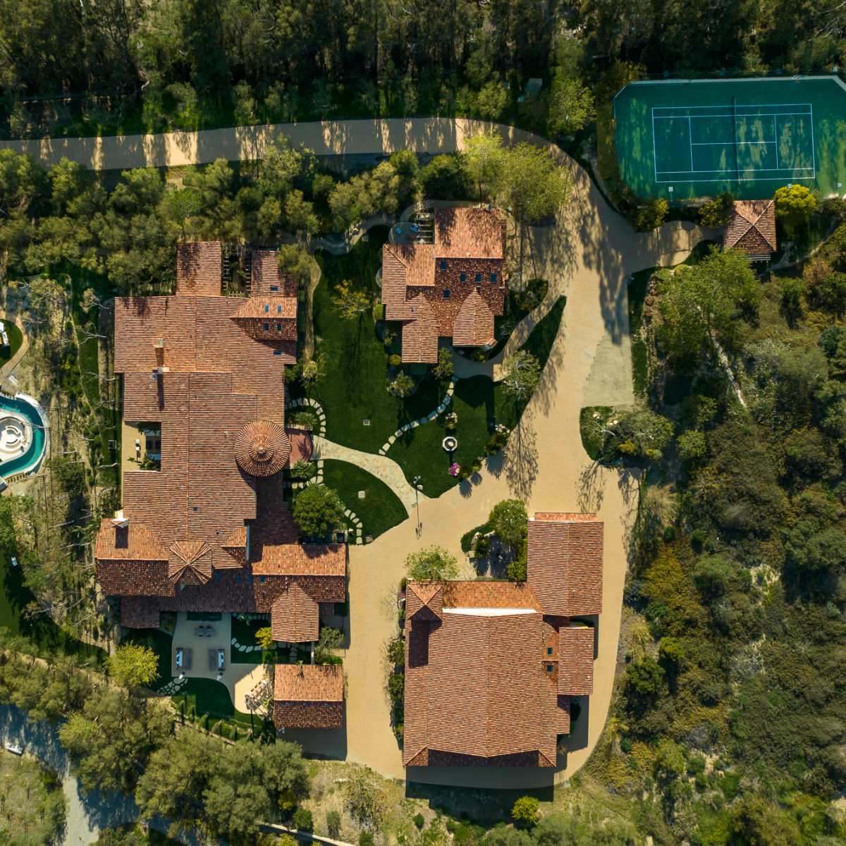 An aerial view of a large estate featuring a main house, guest houses, a swimming pool, and a tennis court set among trees.