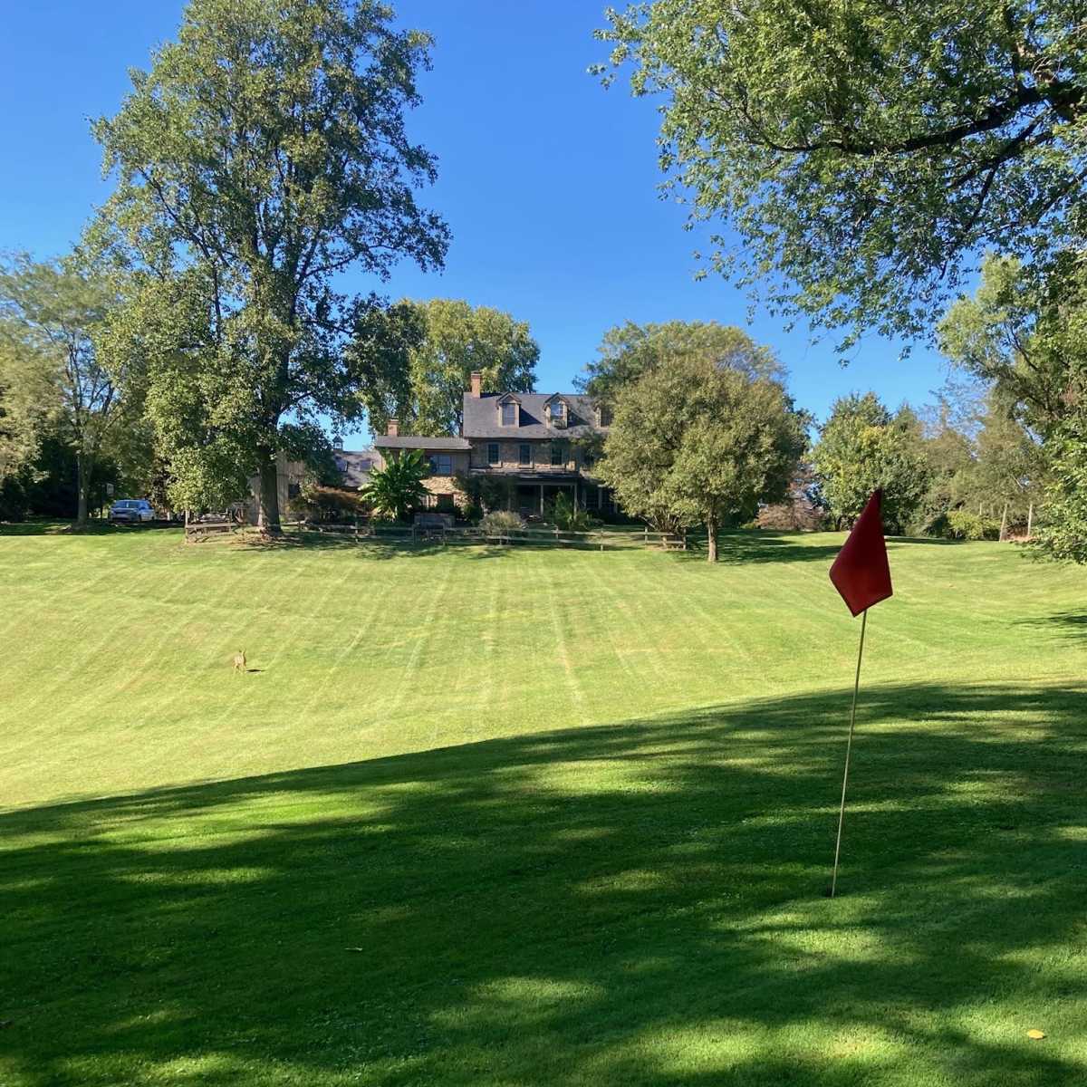 A red flag is located in the foreground of a manicured lawn, leading up to a large house surrounded by trees in the background.