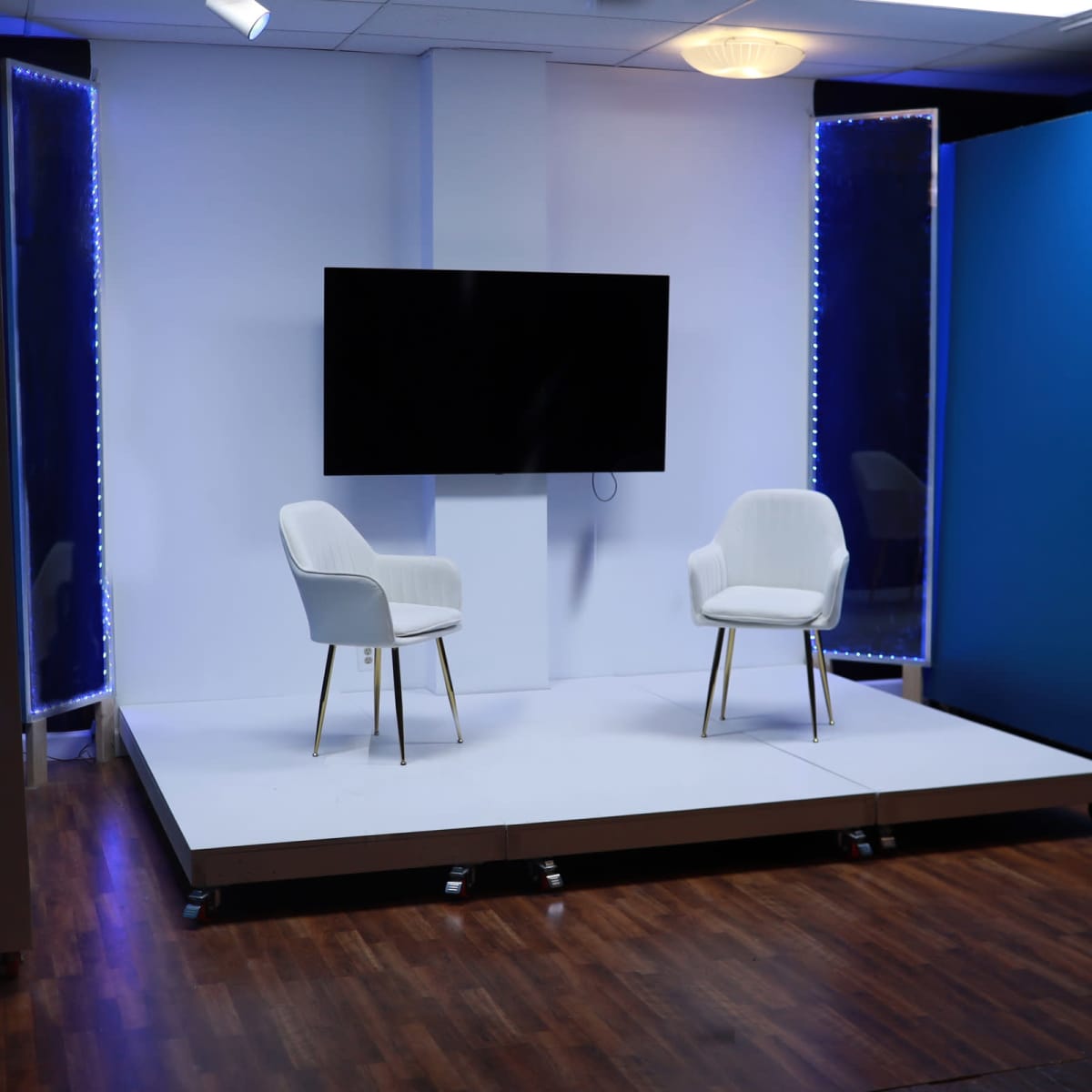 A minimalist studio setup features two white chairs on a raised platform with a large screen and blue backdrop.