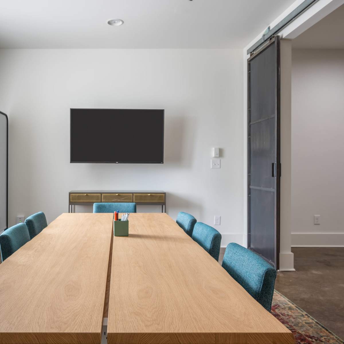 A modern conference room features a long wooden table surrounded by teal chairs, with a wall-mounted television and a large mirror reflected in the space.