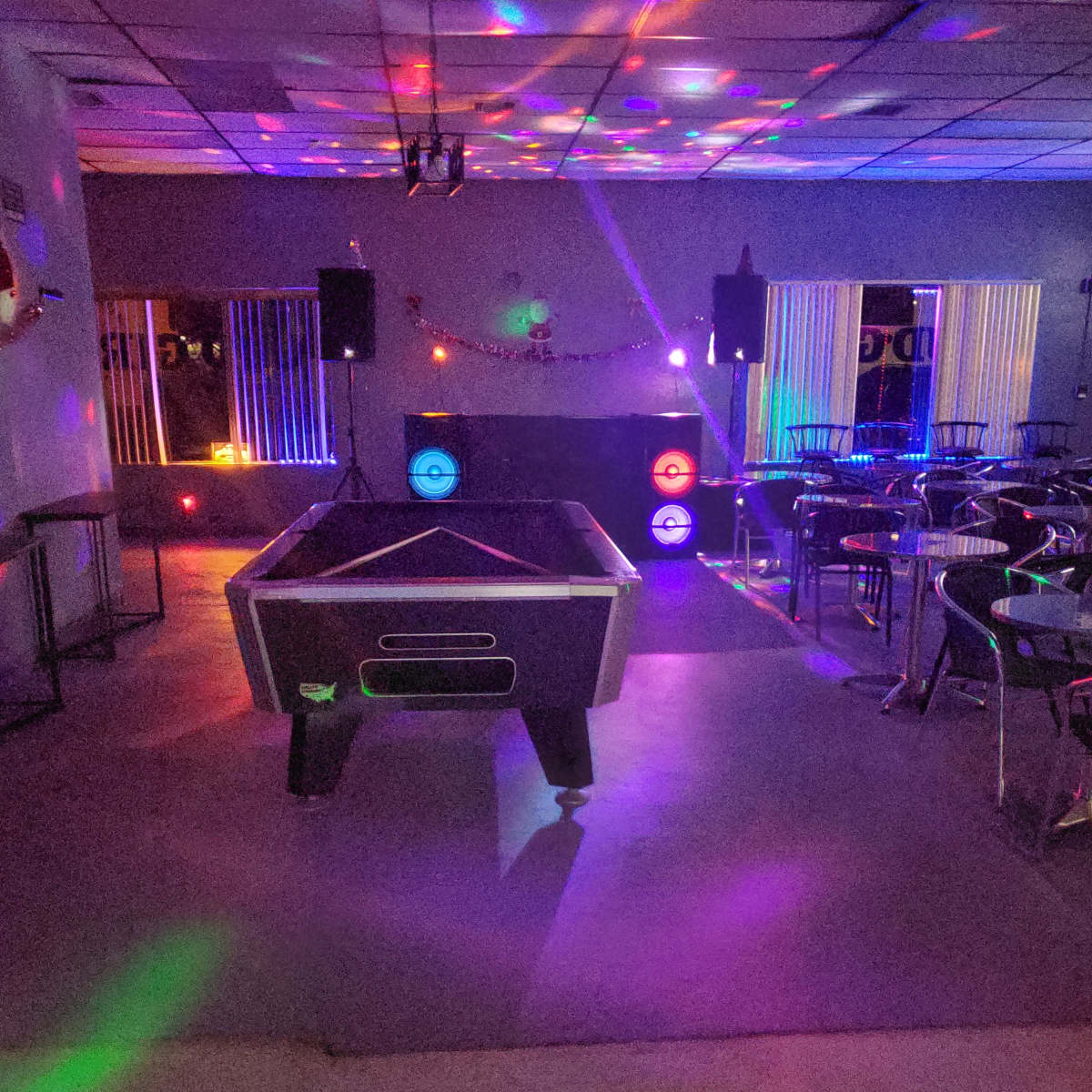 A dimly lit room with a pool table in the center, surrounded by tables and chairs, with colorful lights projecting patterns on the floor and walls.