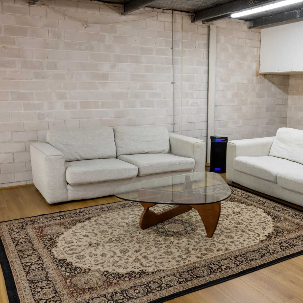 A furnished room with two light-colored sofas, a glass coffee table, and a patterned rug on wooden flooring against a brick wall.