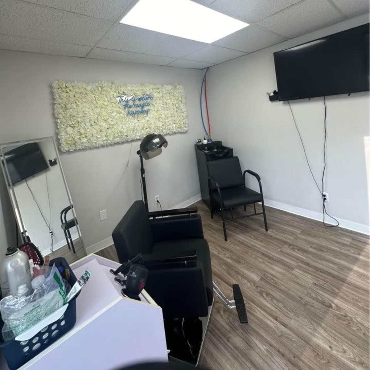 A salon chair positioned in a minimalistic room with a wall of faux flowers, a television mounted on the wall, and wooden flooring.