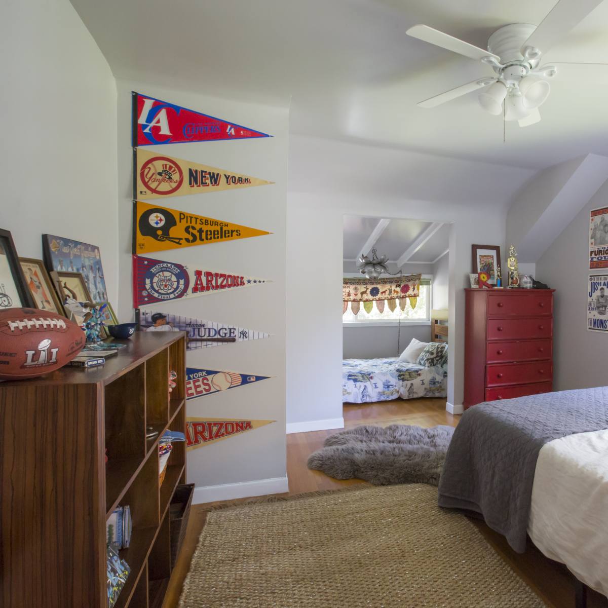 A well-lit bedroom features a bed with gray linens, sports memorabilia displayed on the walls, and a view of another room in the background.
