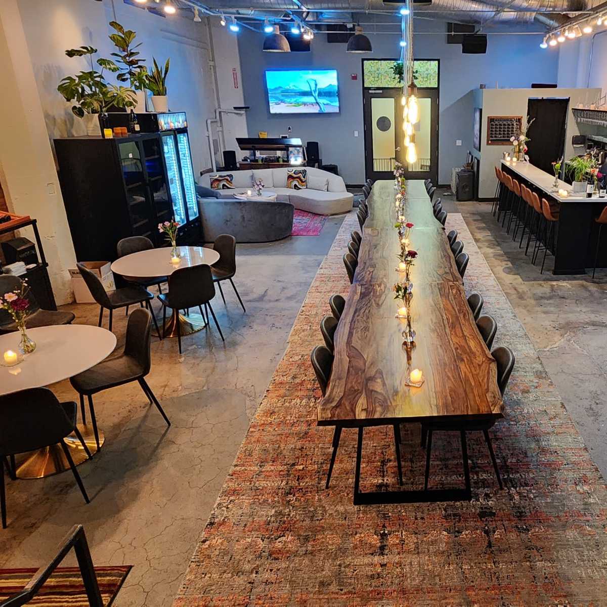 A spacious interior with a long wooden table surrounded by chairs, several smaller round tables, a bar area, and walls lined with bottles of wine.