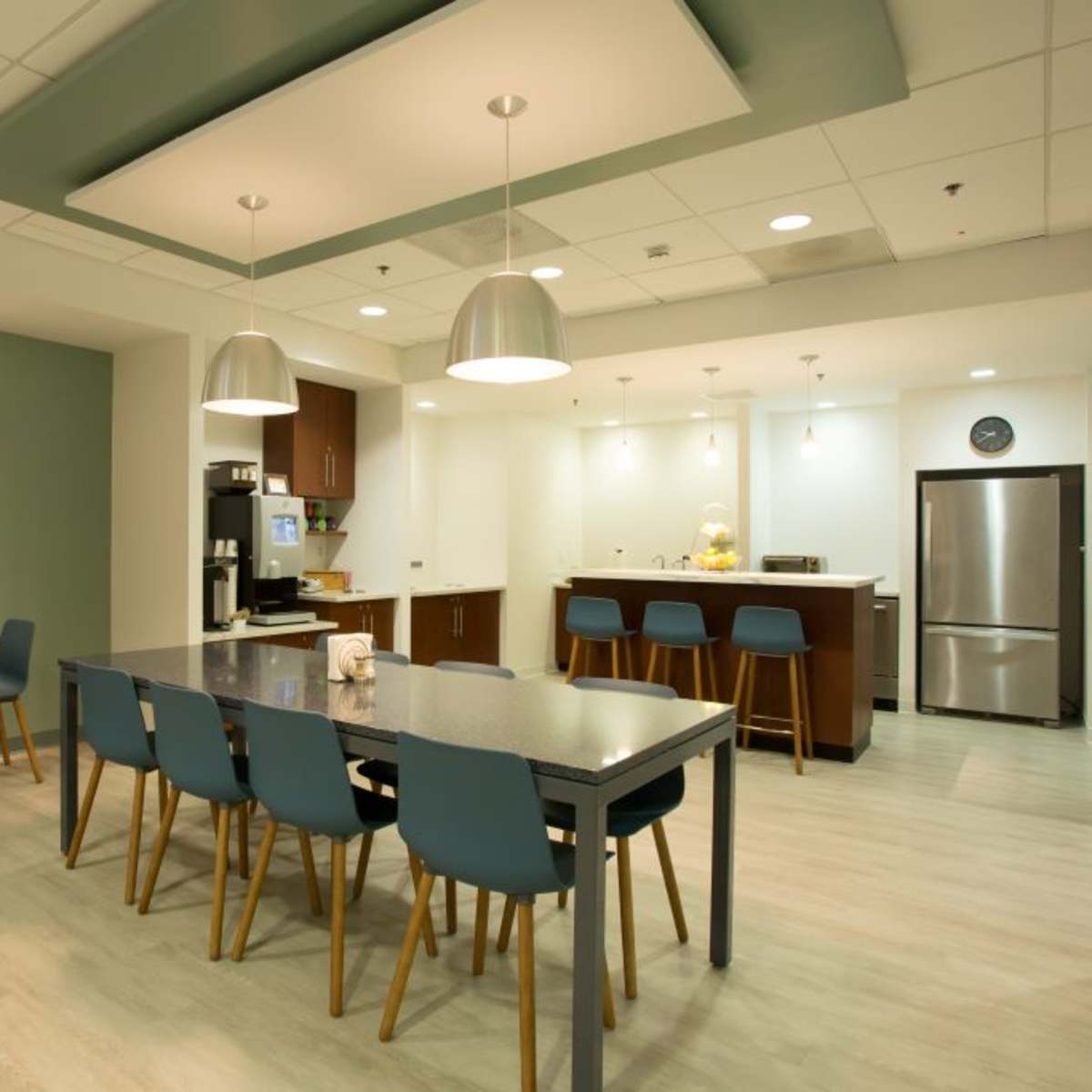 A modern kitchen and dining area features a large table with chairs, a bar counter, and kitchen appliances against a light-colored backdrop.