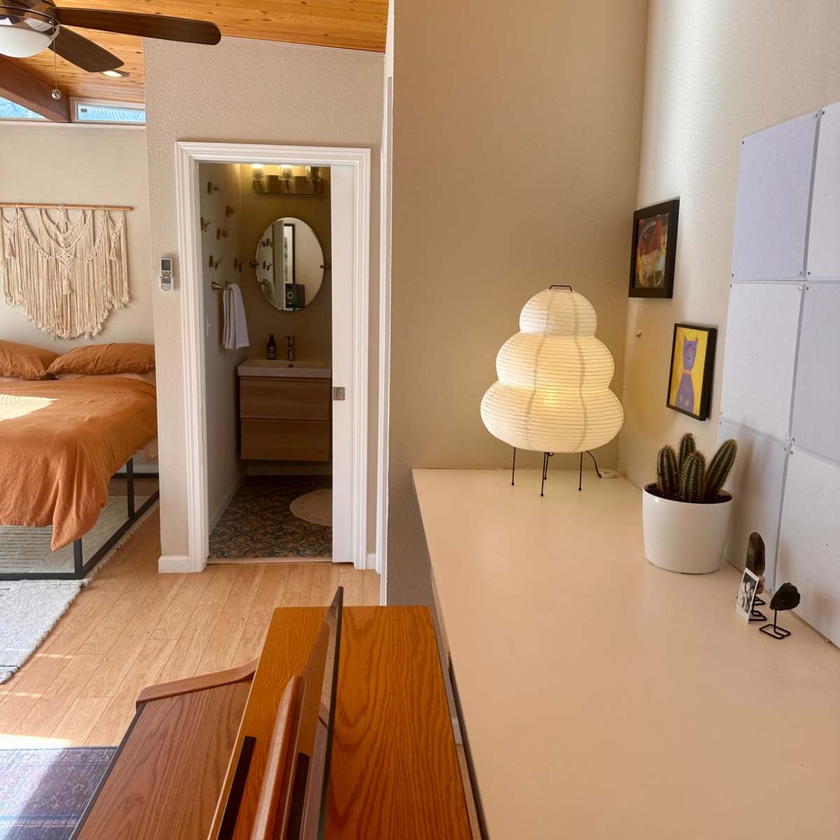 A bright room with a wooden ceiling, featuring a bed in the background, a round mirror, a light fixture on a console table, and a small plant on display.