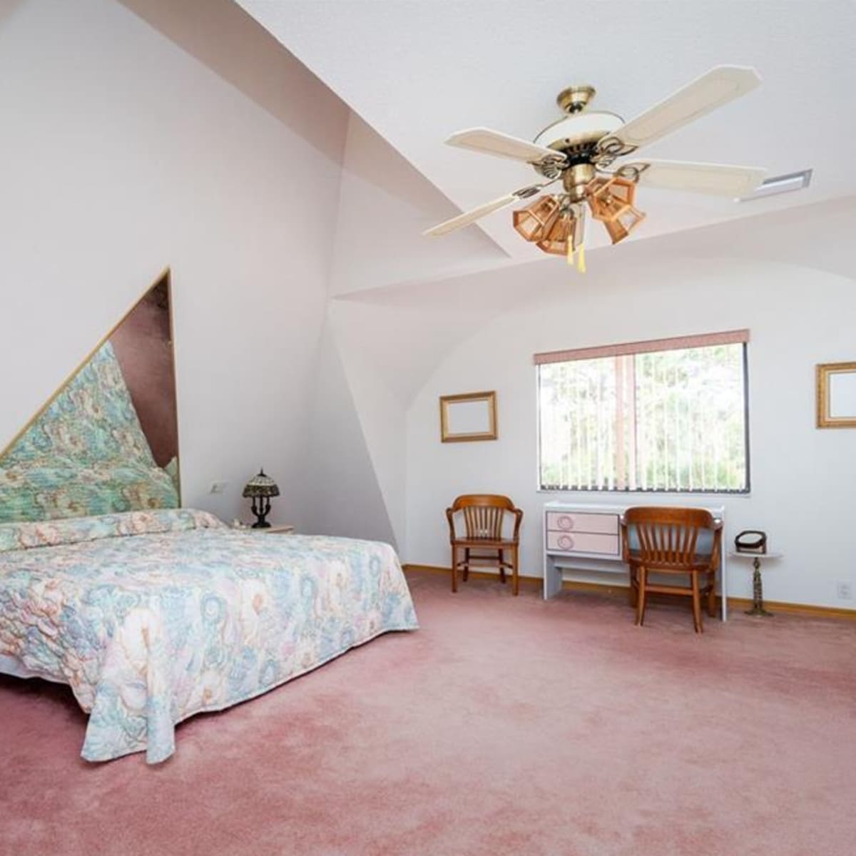 A spacious bedroom features an angled ceiling, a patterned bedspread, wooden furniture, and a window with decorative blinds.