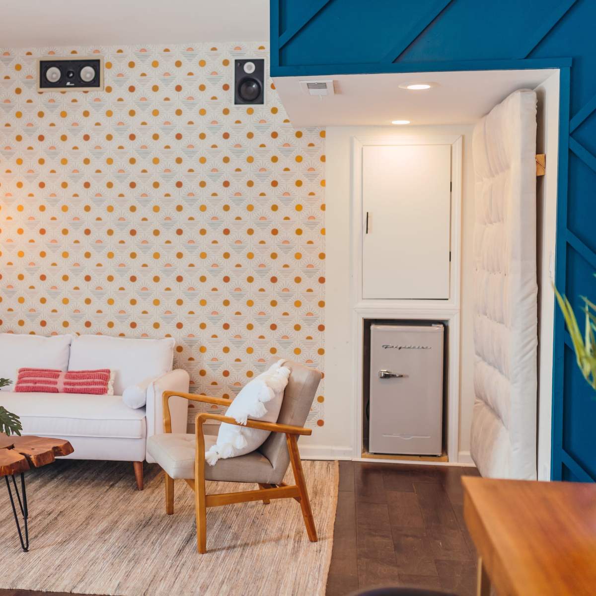 A minimalist living room with a white couch, wooden coffee table, potted plant, and a small refrigerator built into a wall, all arranged against a patterned wallpaper.