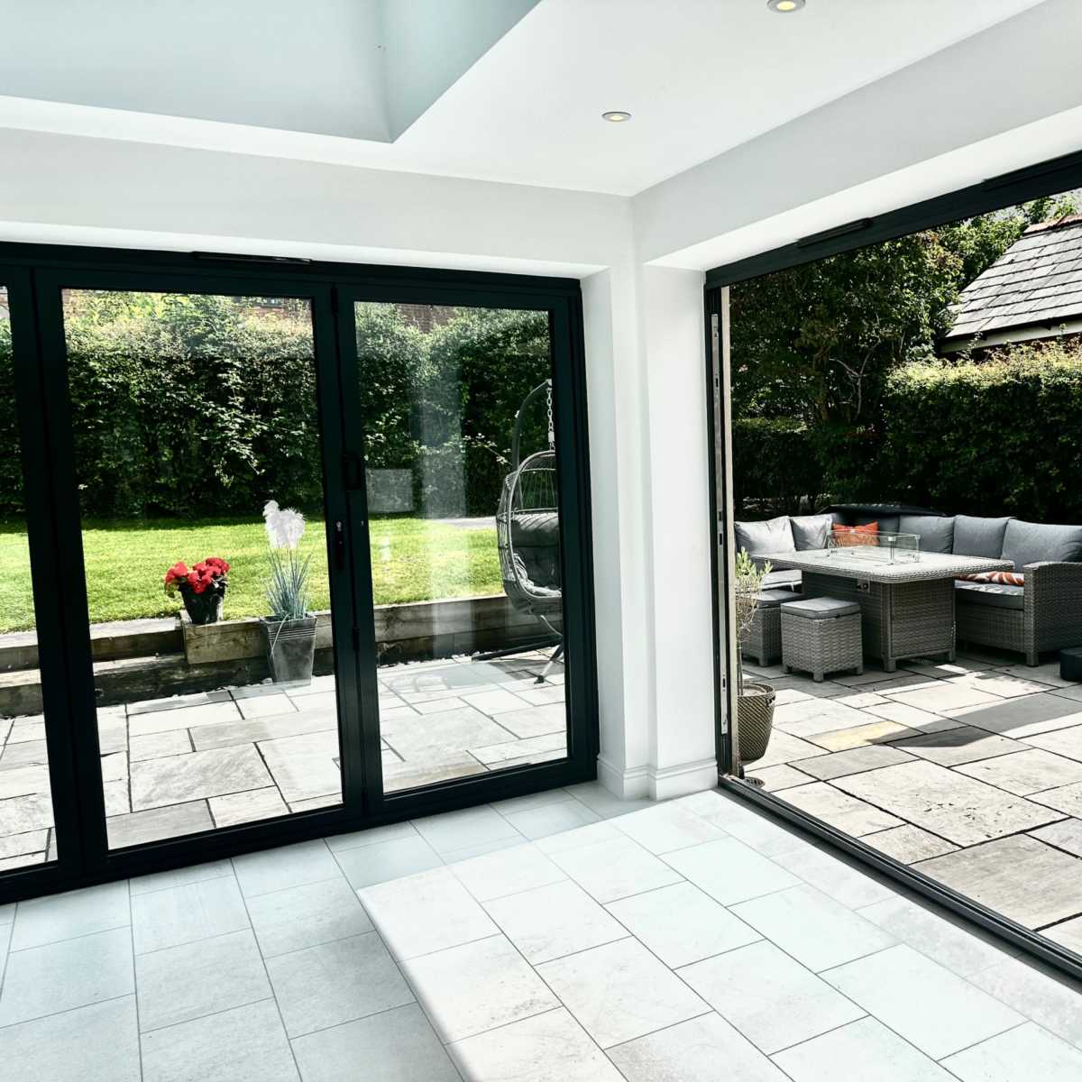 A modern room with large glass doors opening to a patio area featuring outdoor furniture and a well-maintained garden.