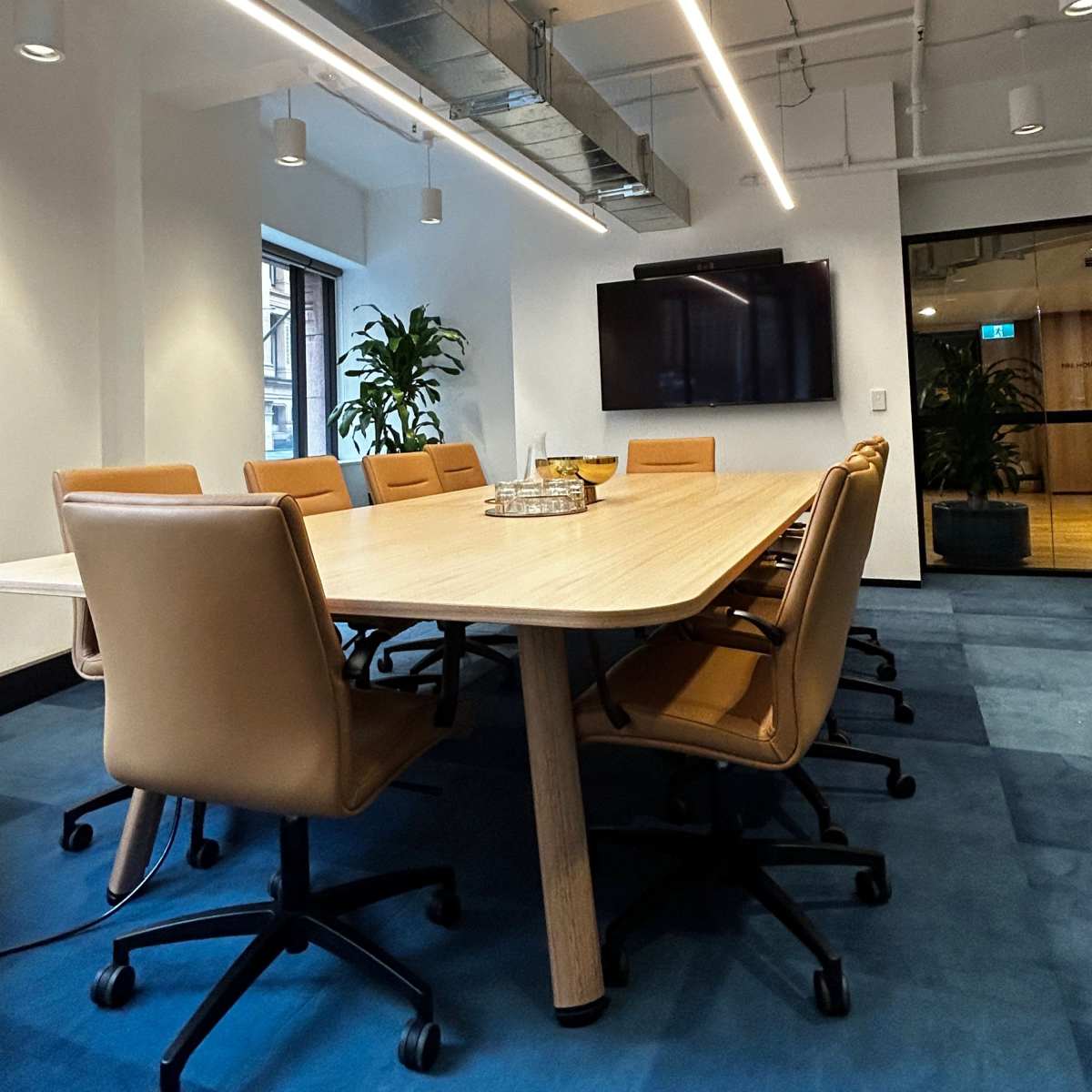 A conference room features a long wooden table surrounded by tan rolling chairs, with modern lighting and a large screen on the wall.