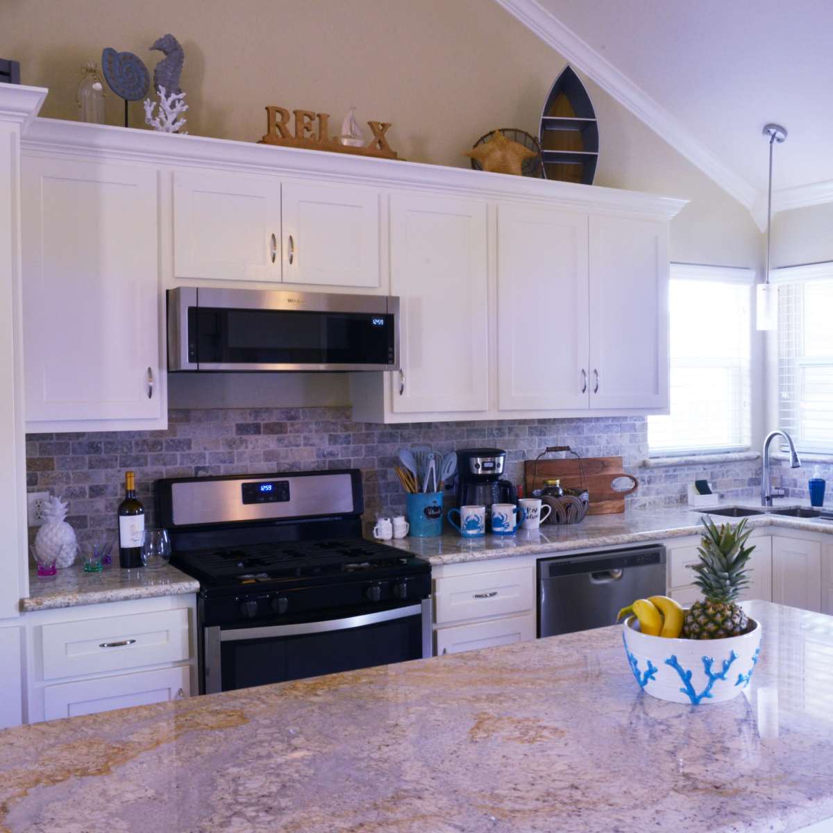 A modern kitchen features white cabinetry, stainless steel appliances, and a granite countertop with a bowl of fruit.