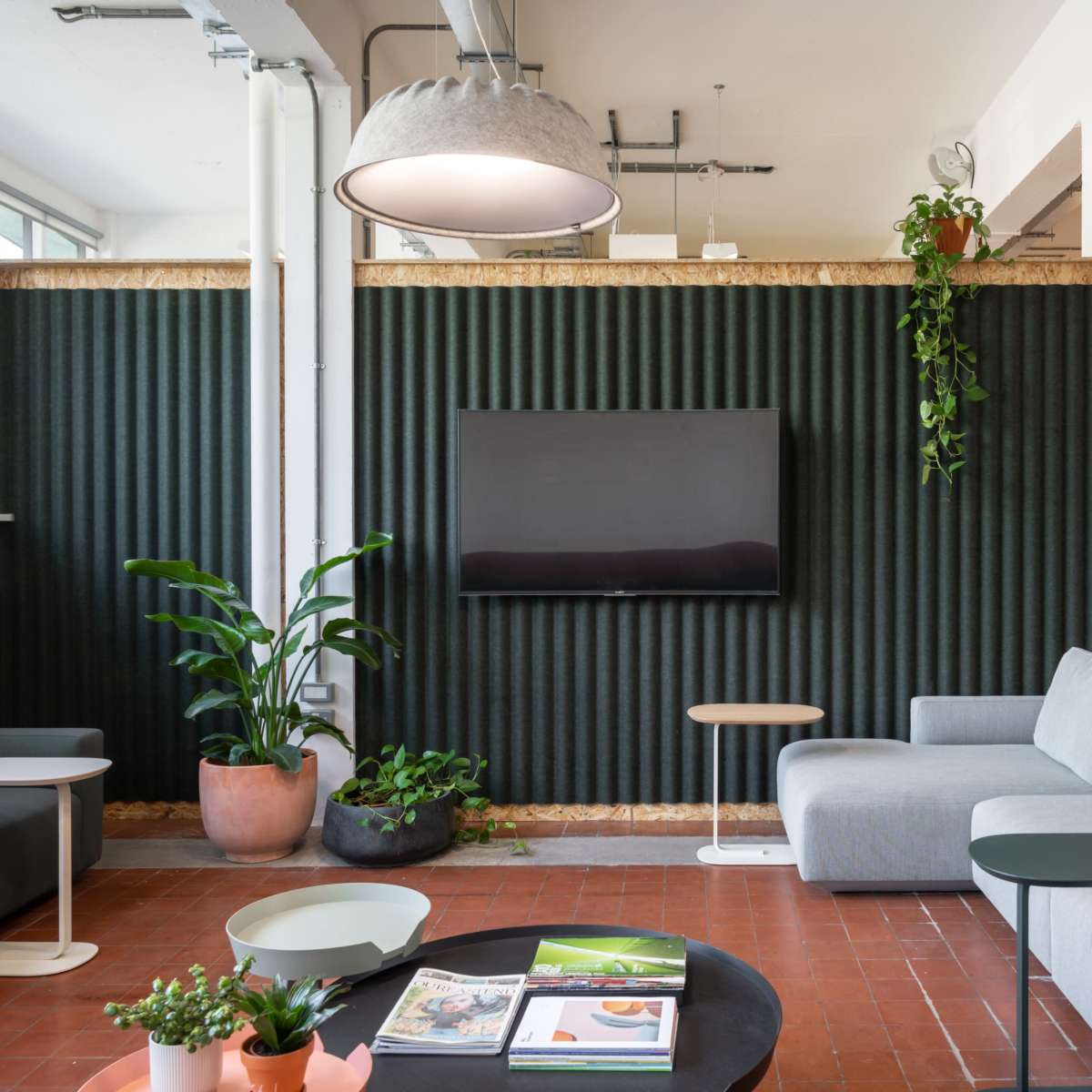 A modern lounge area with gray sofas, a wall-mounted television, and indoor plants against a textured green wall.