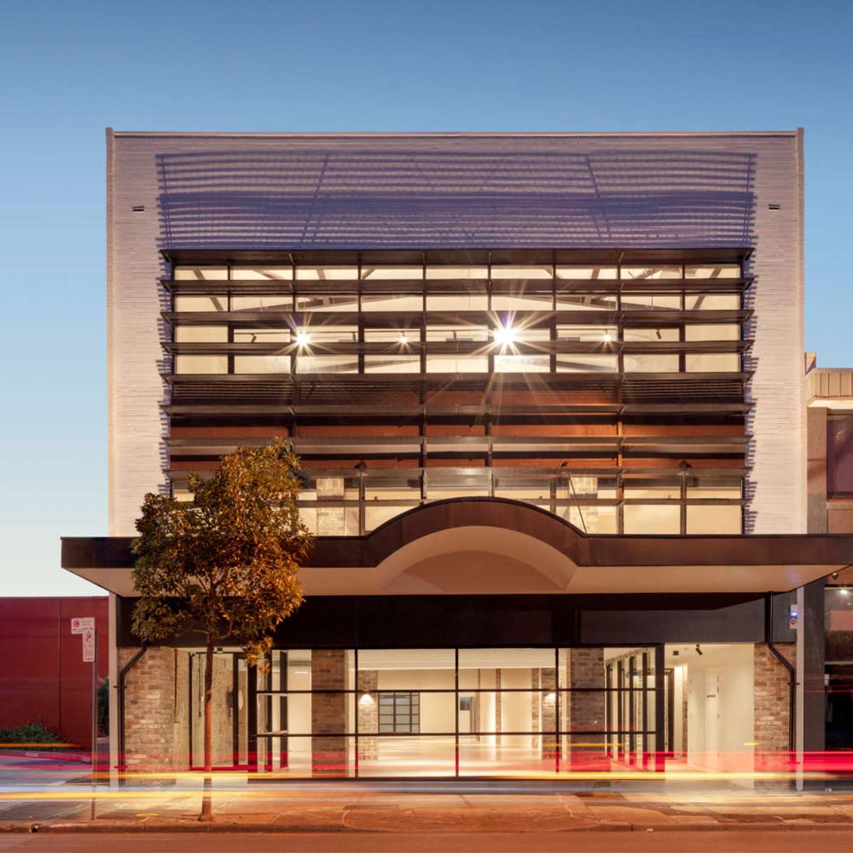 A modern building with large glass windows and a distinctive archway at the entrance, illuminated by evening light.