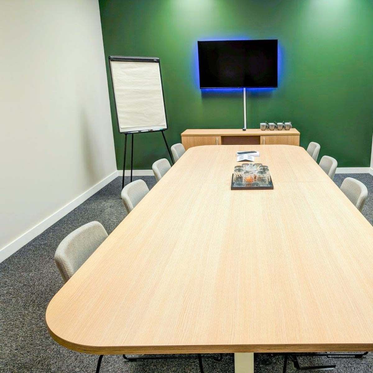 A long, oval conference table is surrounded by ten chairs in a modern meeting room with a television mounted on the wall and a flip chart stand nearby.