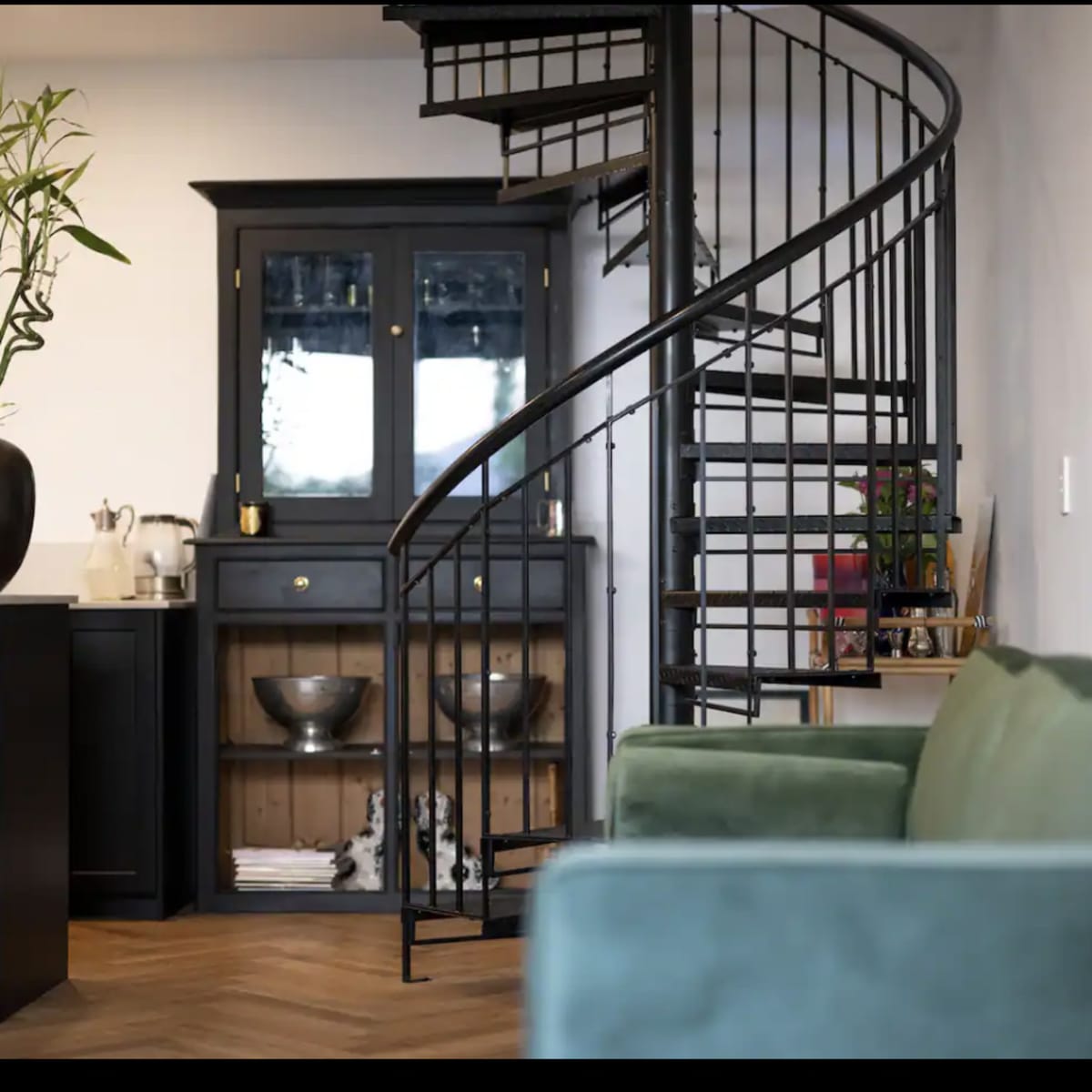 A spiral staircase leading to an upper level in a modern interior, with a green sofa in the foreground and a black cabinet against the wall.