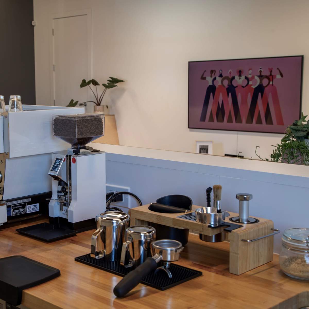 A modern coffee setup featuring an espresso machine, grinder, and various coffee brewing tools on a wooden countertop, with a colorful artwork displayed on the wall behind.