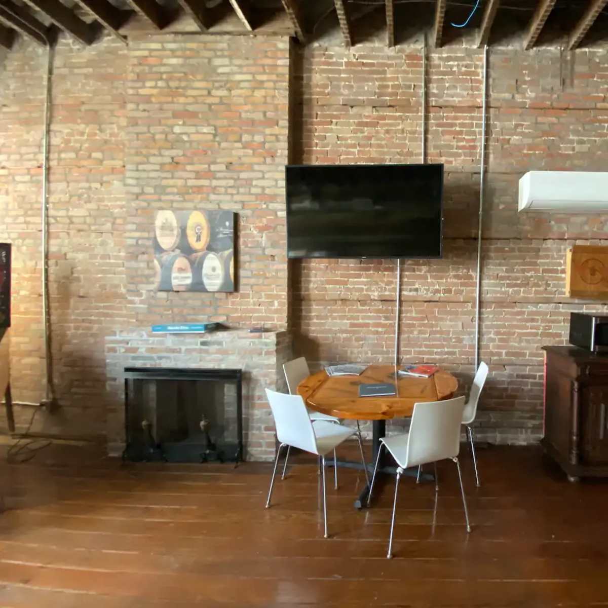 A room with exposed brick walls, featuring a round wooden table with four white chairs, a wall-mounted TV, and a wooden cabinet beside a fireplace.