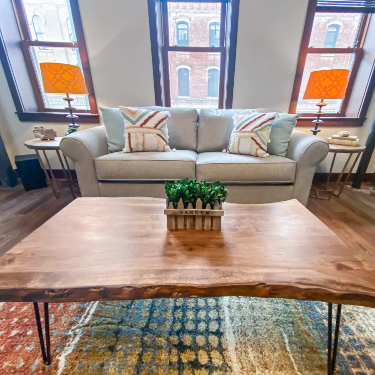 A light gray sofa with decorative pillows is placed in a room featuring a wooden coffee table and two lamps with orange shades beside large windows.