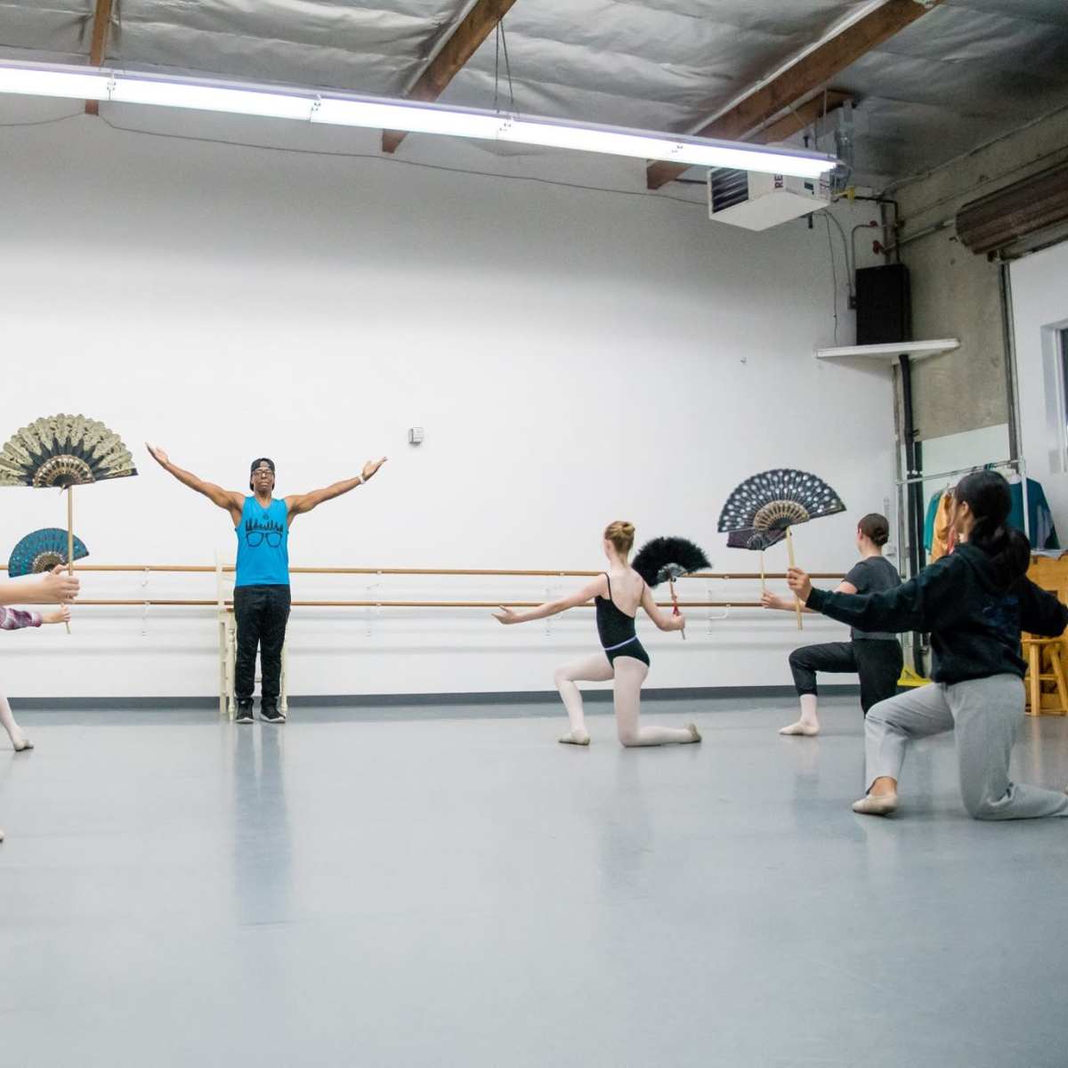 A group of dancers practices in a studio while holding fans, with an instructor positioned in front, raising his arms.