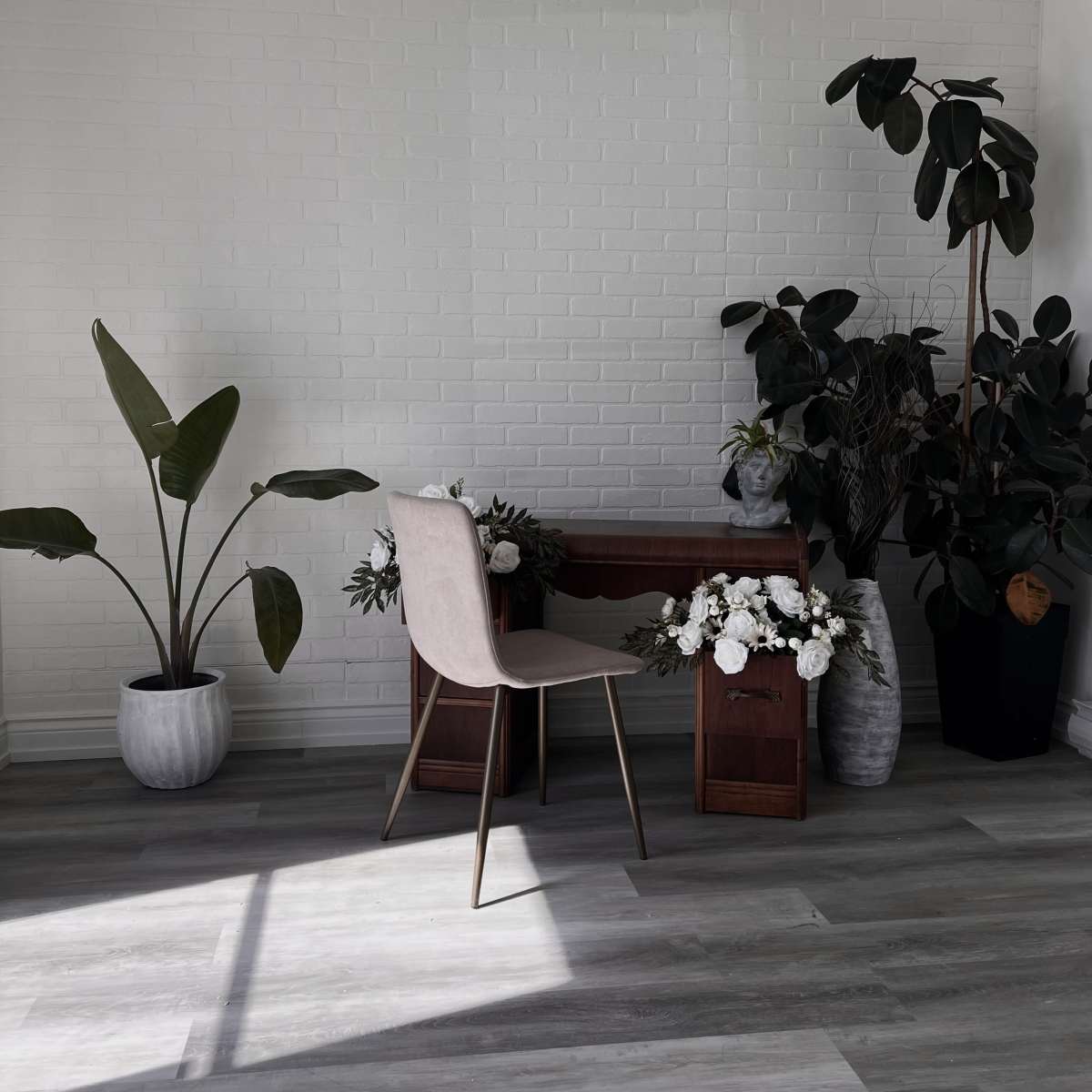 A minimalist workspace features a chair and desk surrounded by plants and floral arrangements against a white brick wall.