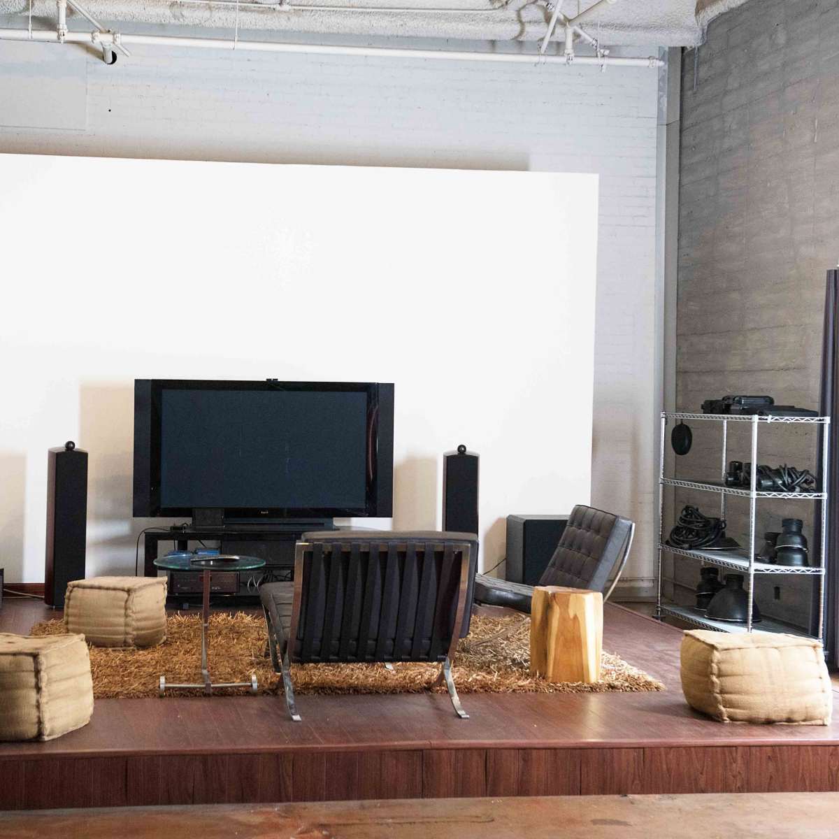 A modern living room setup features a large television on a white wall, surrounded by minimalist furniture, including two black chairs, a coffee table, and several seating poufs on a wooden floor.
