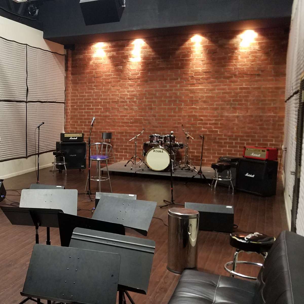 A music rehearsal studio featuring a drum set, amplifiers, and several music stands arranged in the space.