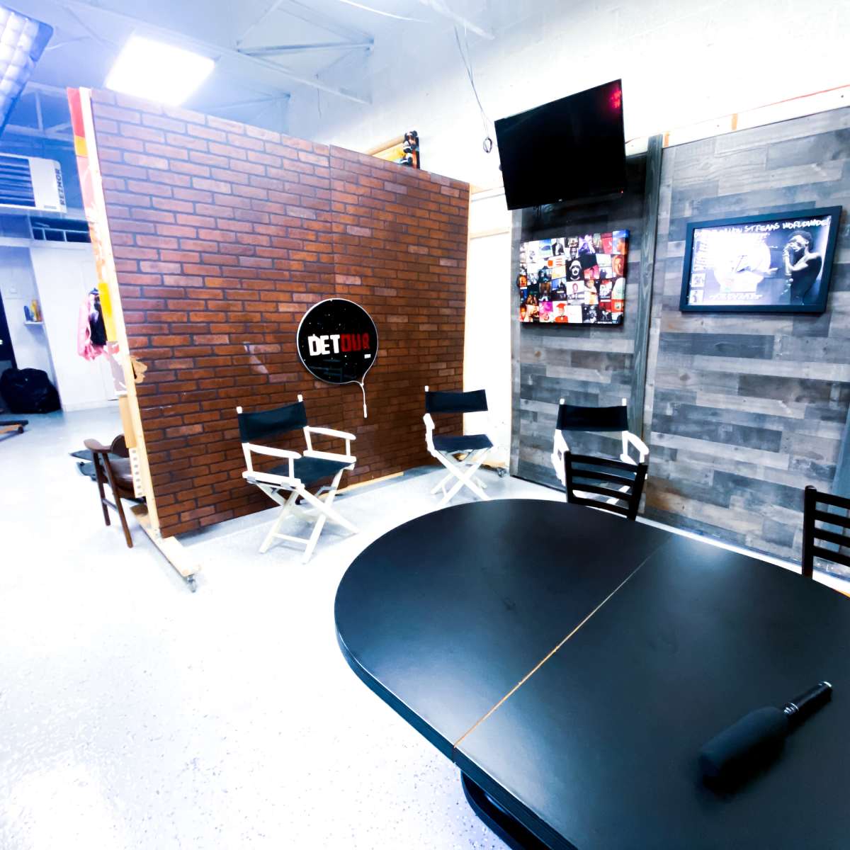 A studio space with a brick wall backdrop, a black circular table, director's chairs, and a television displaying photos and graphics on the walls.
