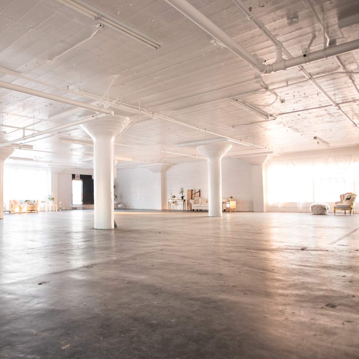 A spacious, empty industrial loft with large windows, concrete floors, and several white columns.