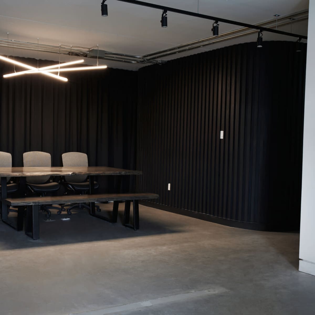 A conference room features a dark wall with three chairs and a long table under modern light fixtures.