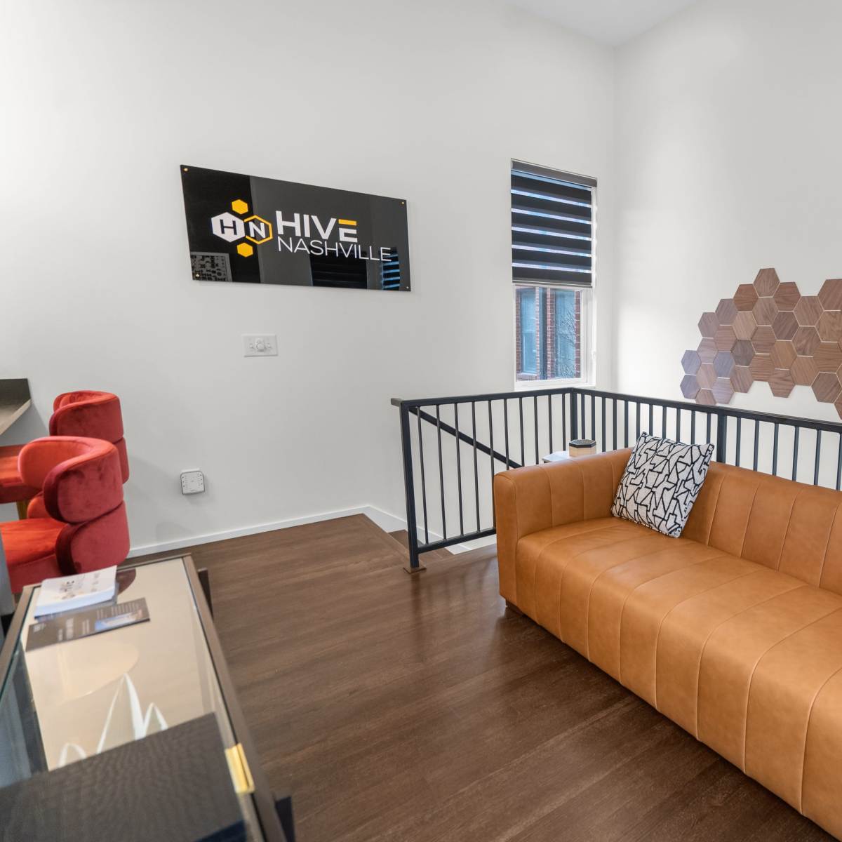 A modern lounge area featuring a tan leather couch with patterned cushions, a black railing, and a decorative hexagonal wall art.