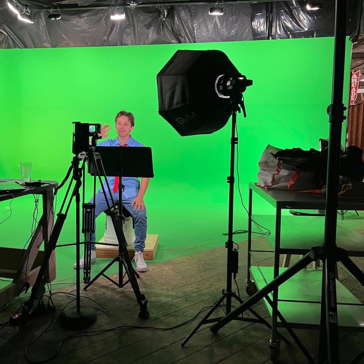 A person sits on a platform in front of a green screen, surrounded by lights and a camera setup.