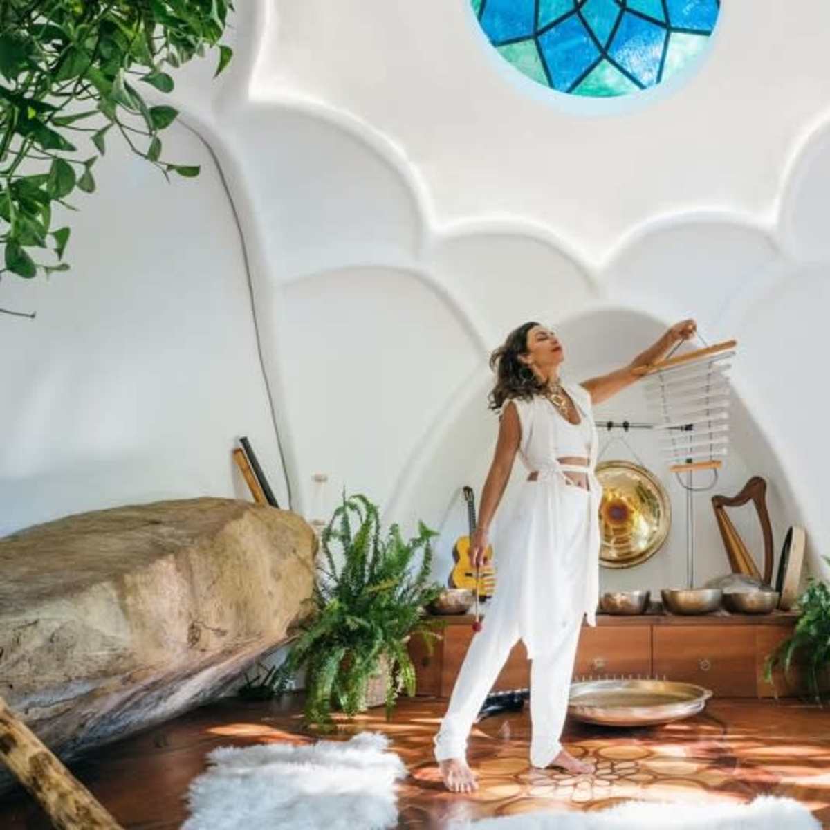 A woman in white clothing stands in a brightly lit, uniquely shaped room, holding a wind chime, surrounded by plants and musical instruments.